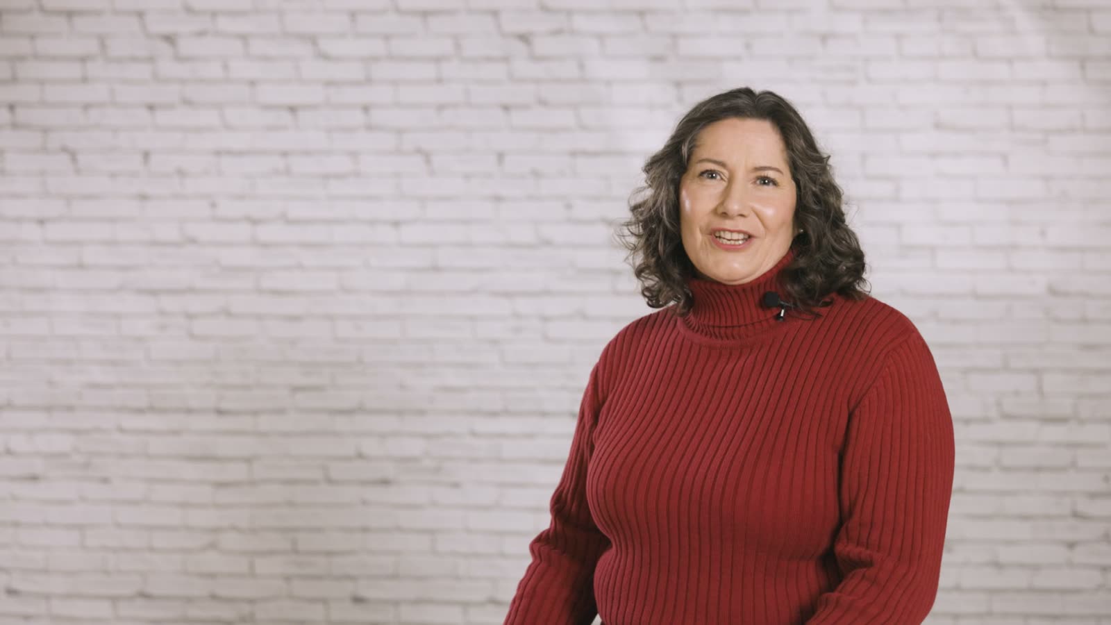 A woman in a red polo-neck jumper smiles into the camera in front of a white brick wall