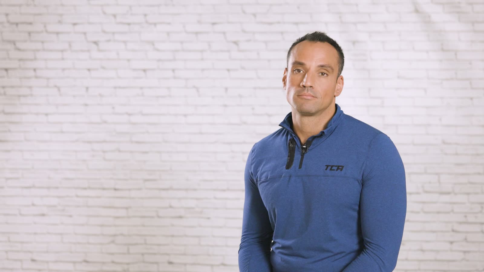 A man in a blue sports top speaks into the camera in front of a white brick wall