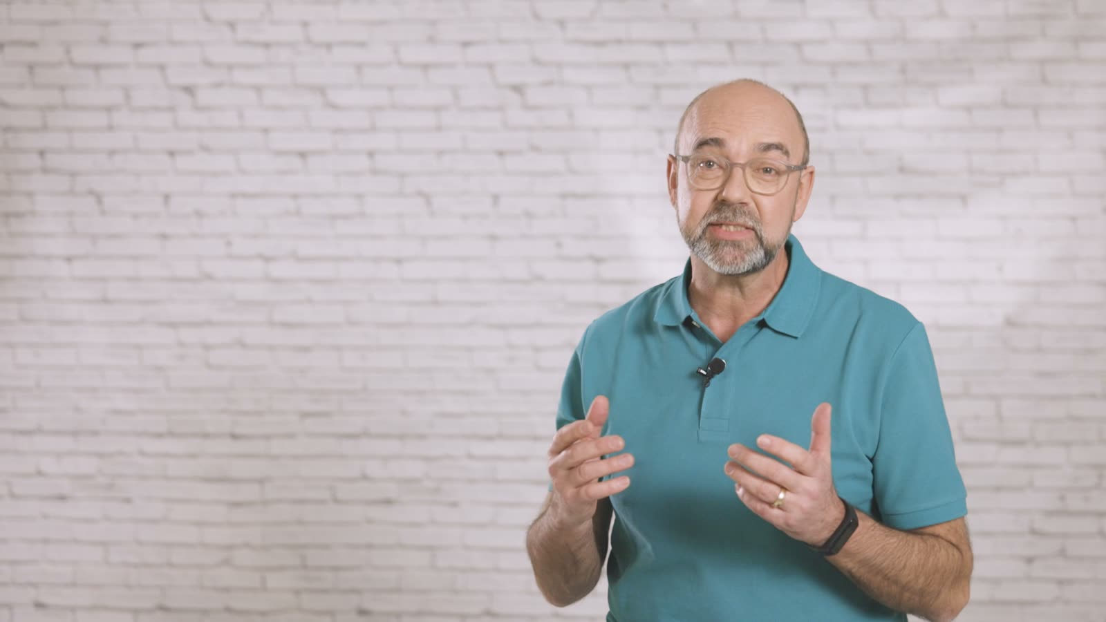 A man with glasses in a teal polo shirt speaks into the camera in front of a white brick wall