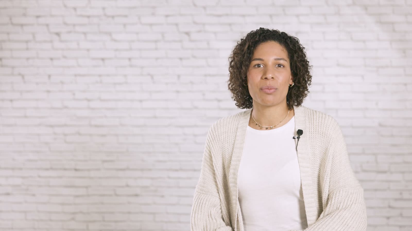 A woman with curly hair speaks into the camera in front of a white-painted brick wall