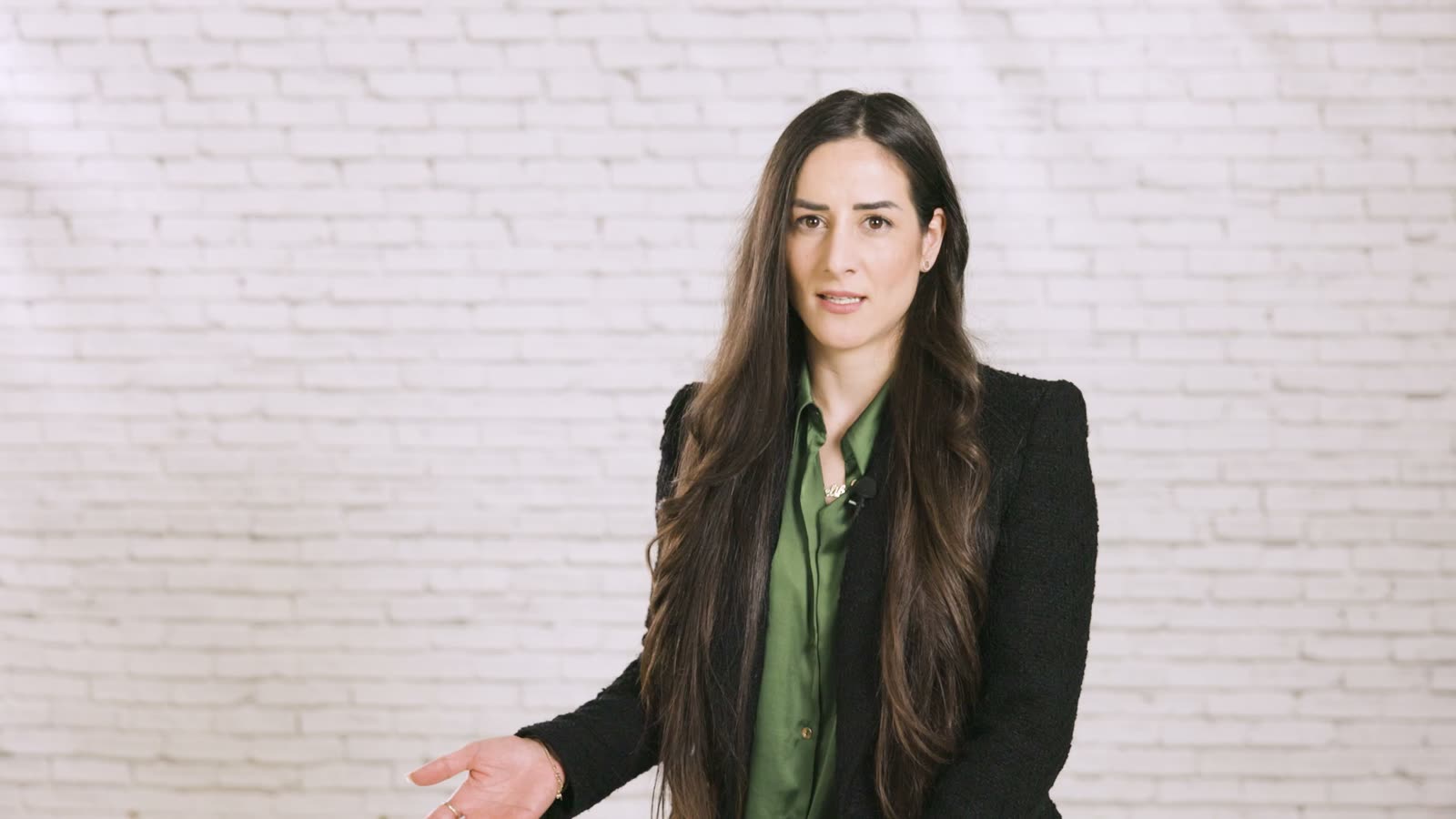 A woman in a dark blazer over a green blouse speaks into the camera in front of a white brick wall