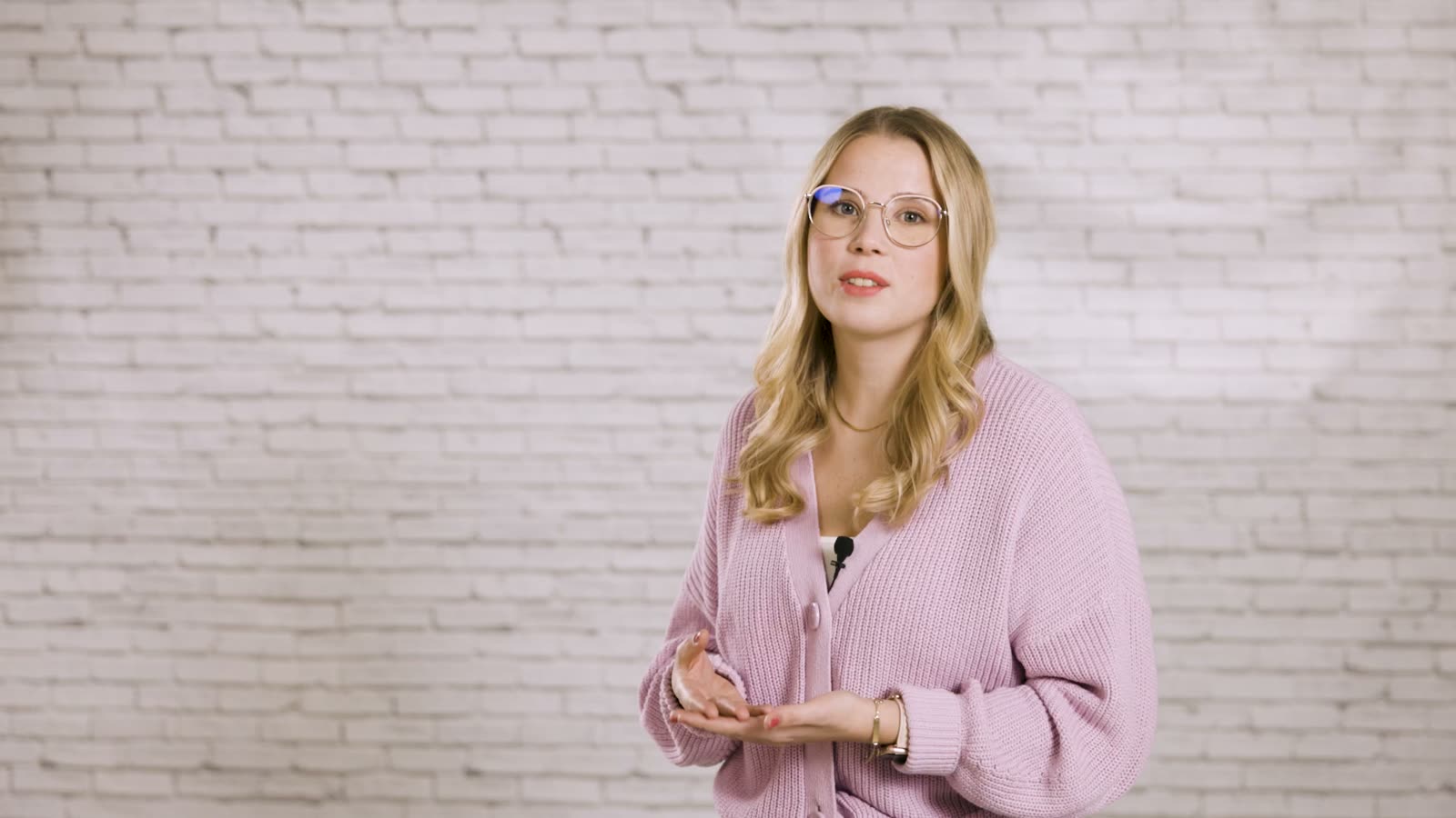 A young woman with glasses and a pink cardigan speaks into the camera in front of a white brick wall