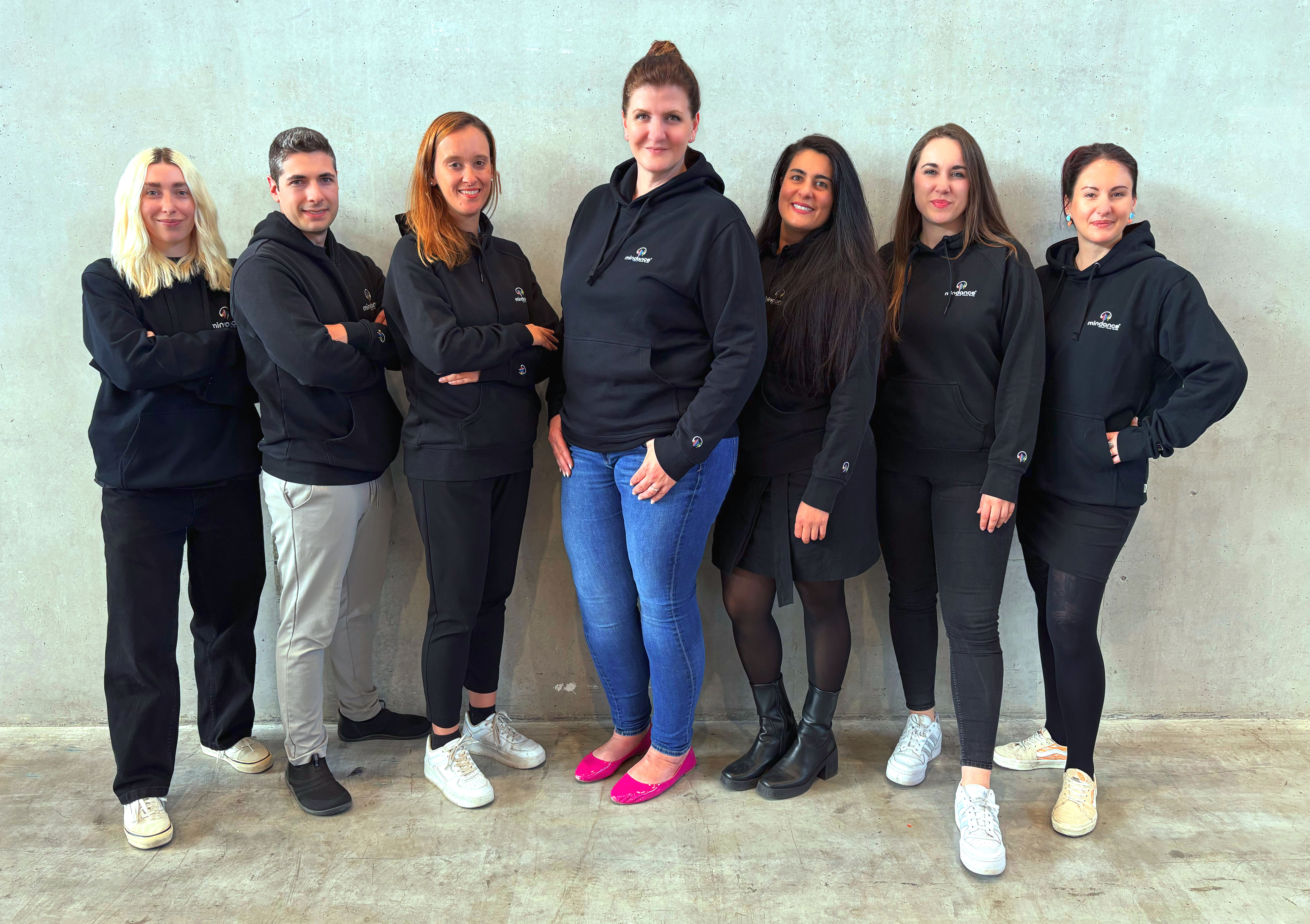 Das Mindance Team, sieben Personen nebeneinander vor einer hellen Betonwand