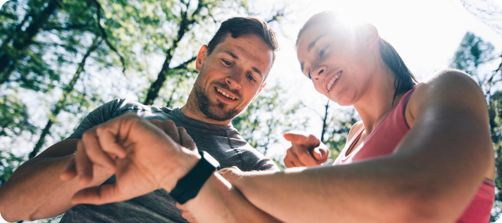Two athletes in a forest smile as they look at a sports watch together