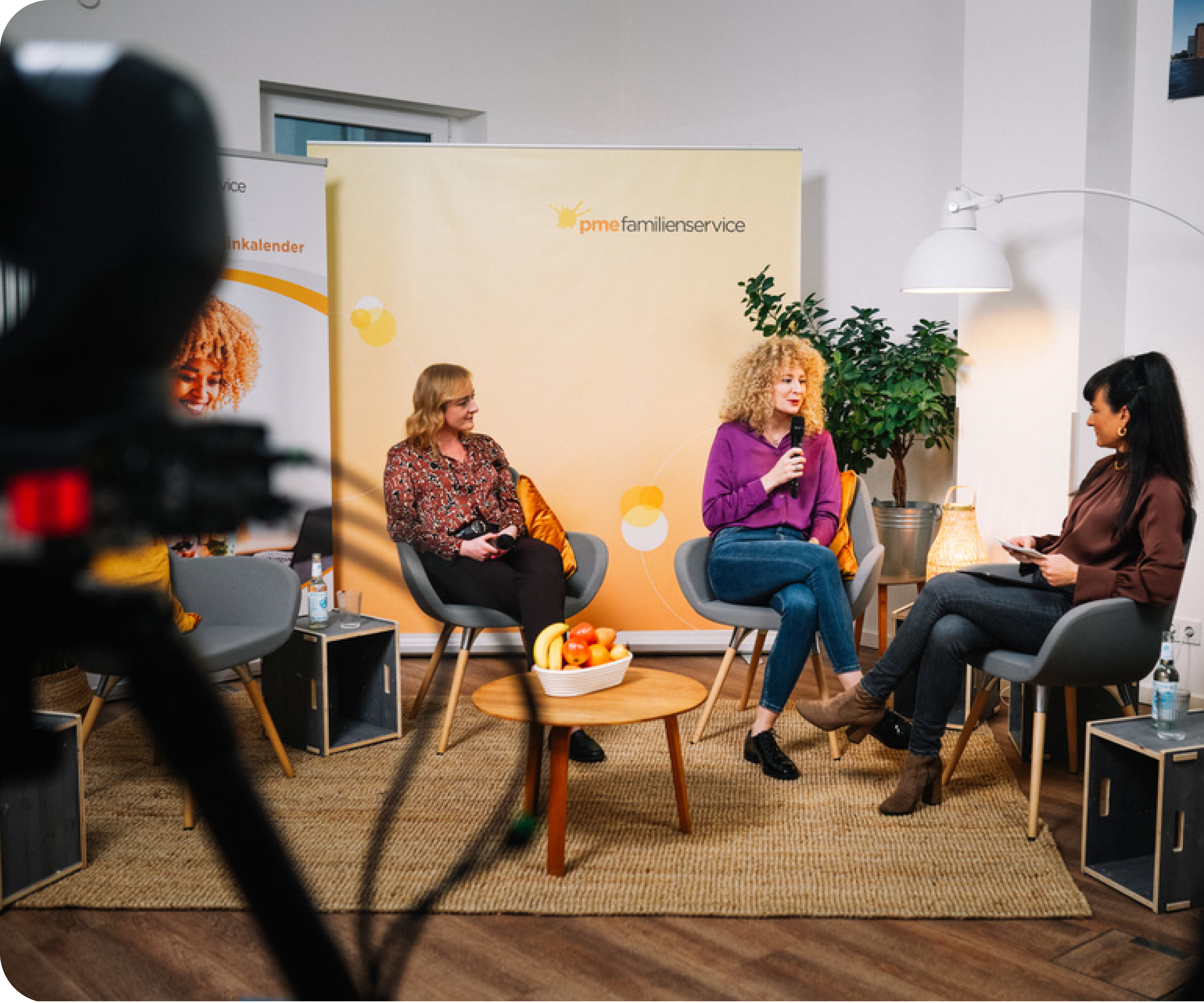 Three women sit talking in a studio, a pme Familienservice roll-up behind them and a camera in the foreground
